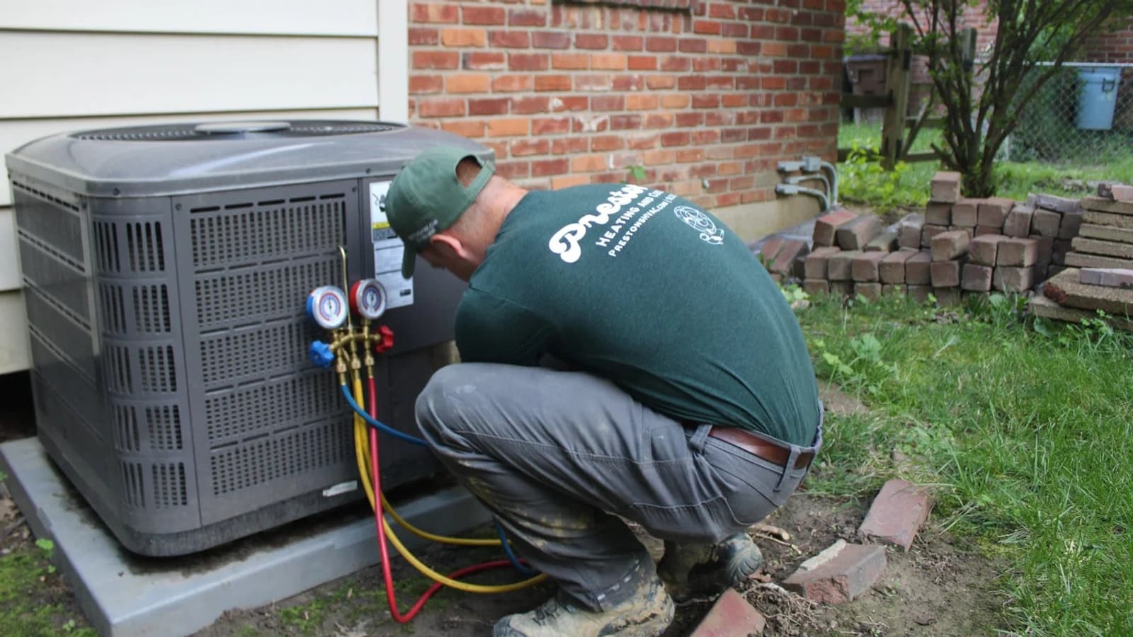 HVAC technician in a green cap crouching beside an outdoor air conditioning condenser, connecting refrigerant gauges